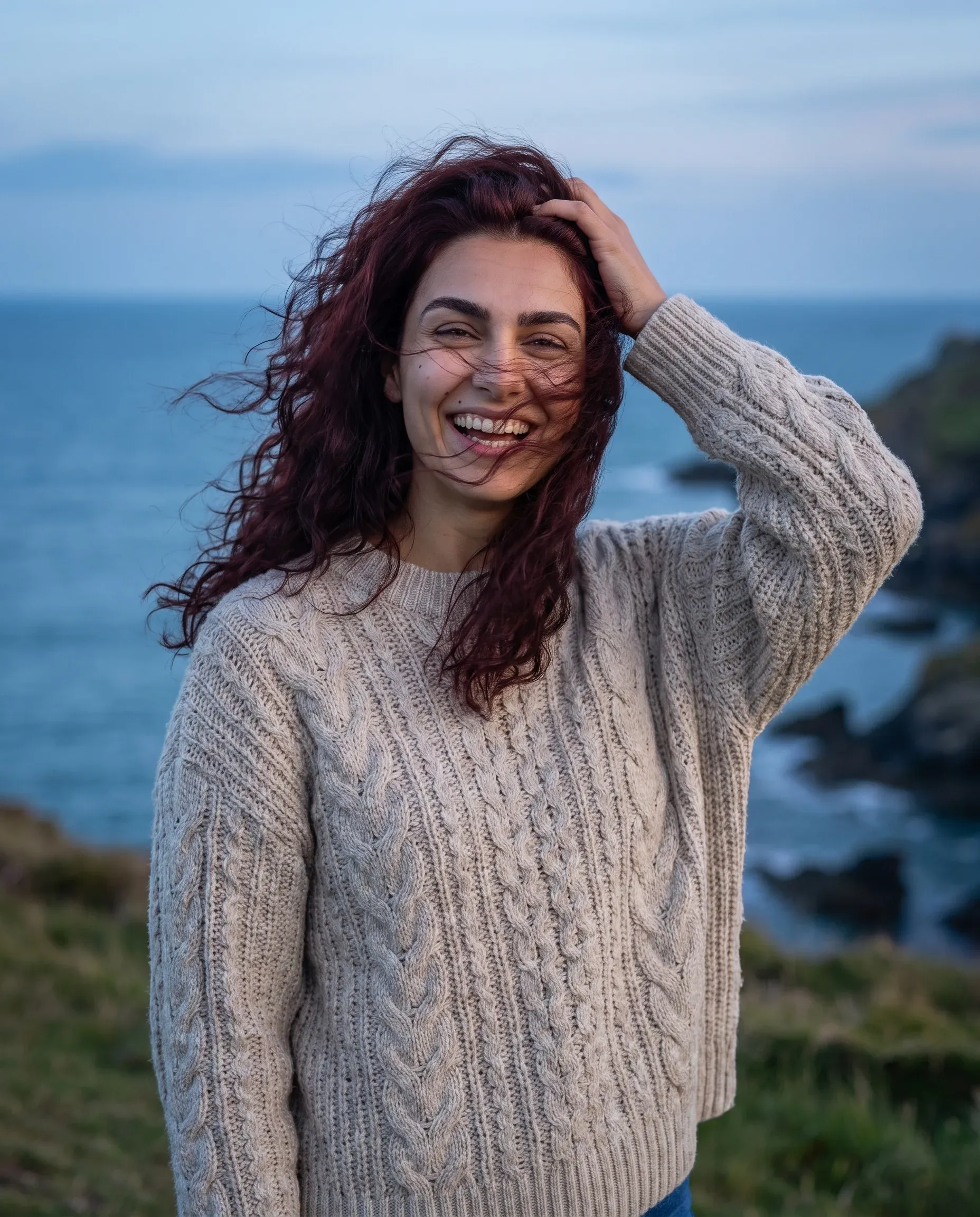 The same character on a coastal headland at dusk in a cable-knit sweater, laughing with her hair blowing across her face