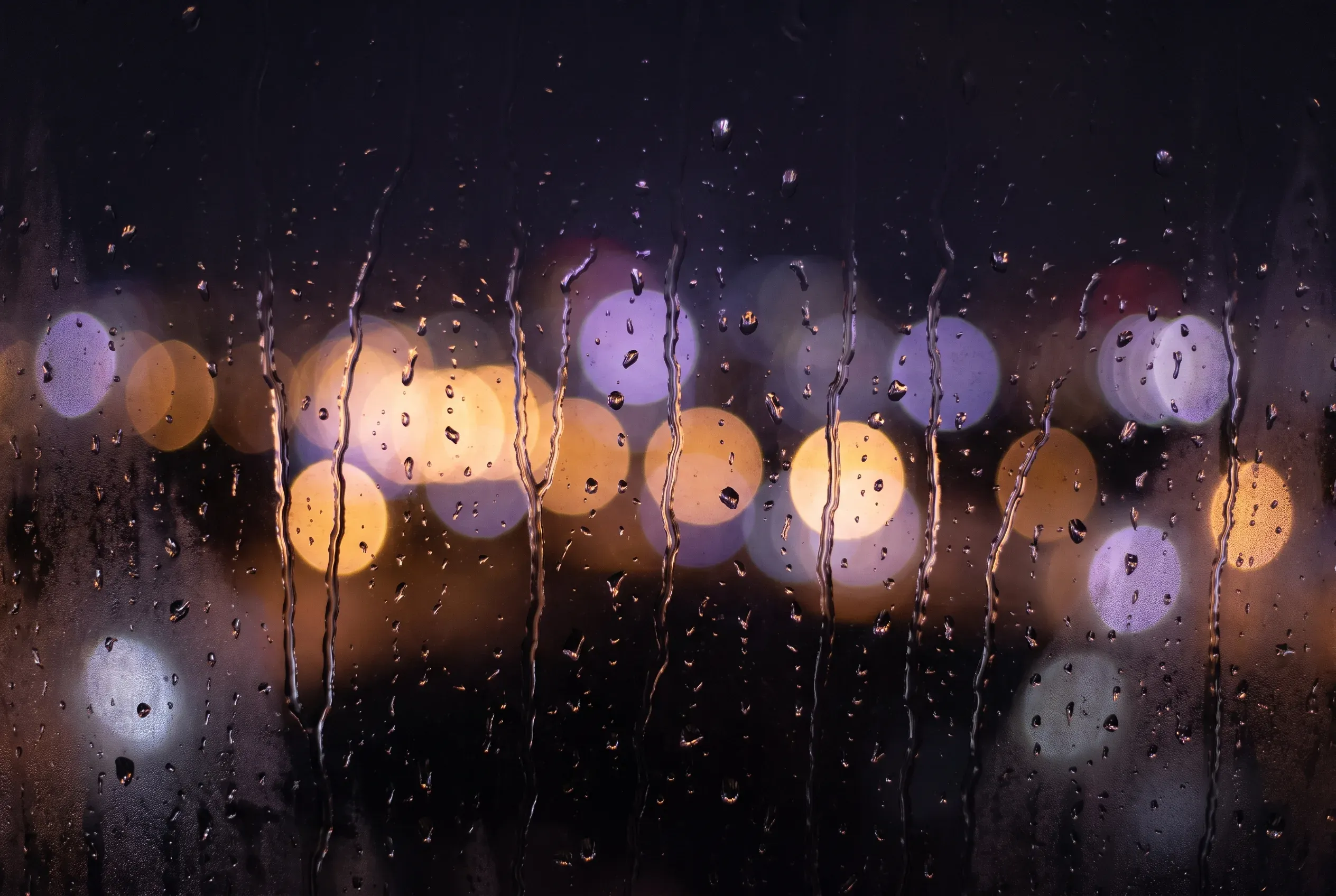 Rain running down a dark window with city lights behind it thrown into soft violet and amber bokeh