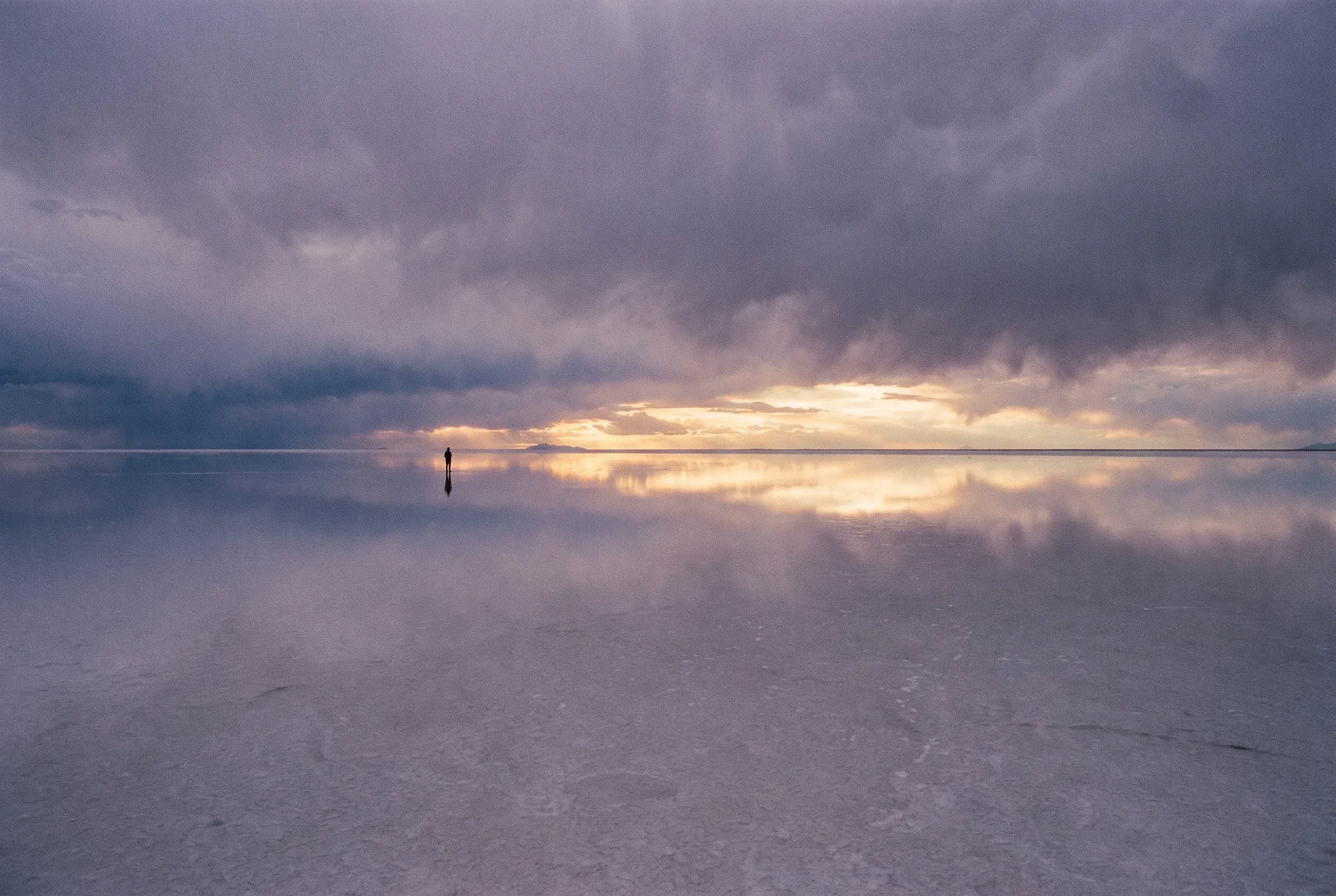 A mirrored desert salt flat reflecting a towering storm sky, with a lone distant figure for scale